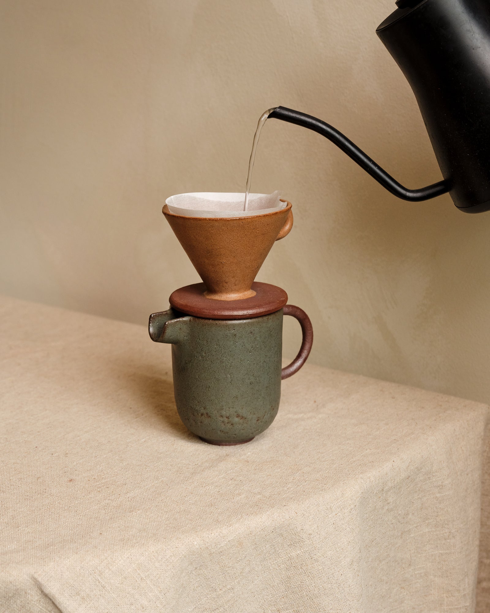 Ceramic coffee maker with a ceramic filter being filled with hot water on a beige surface.