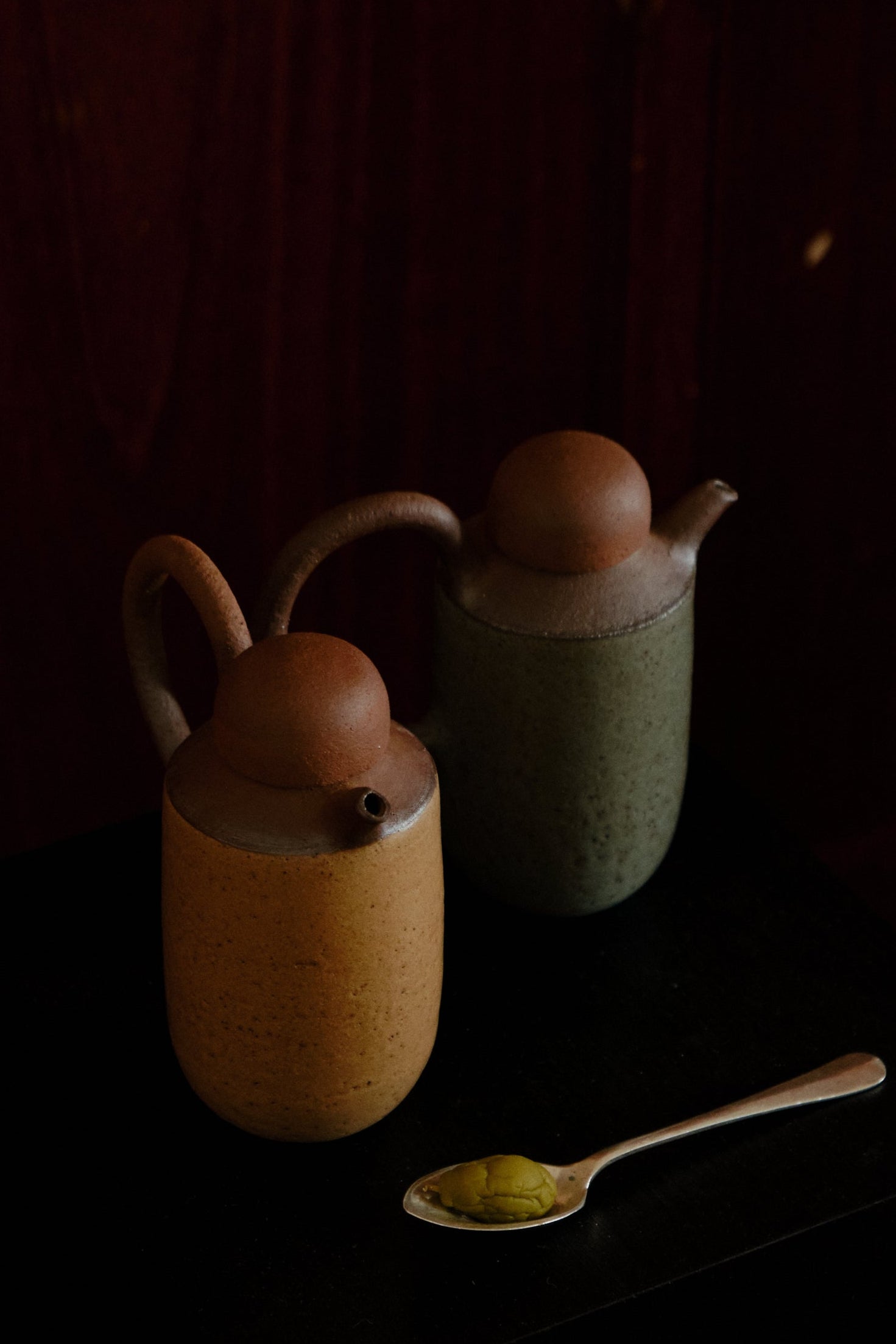 A ceramic olive oil bottle with a rustic texture, displayed in a dark setting, with a wooden spoon beside it.