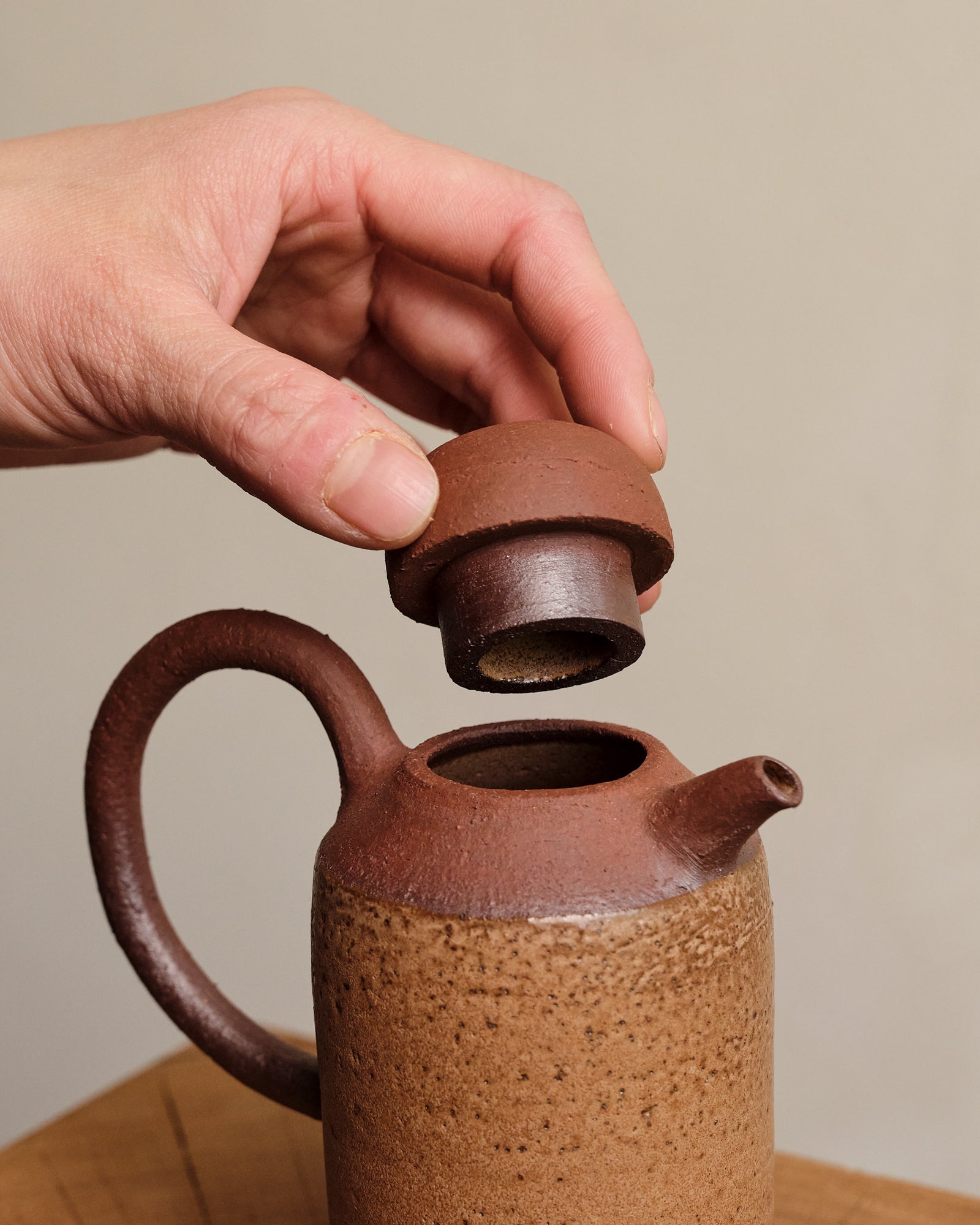 Person opening a brown ceramic bottle lid with a neutral background