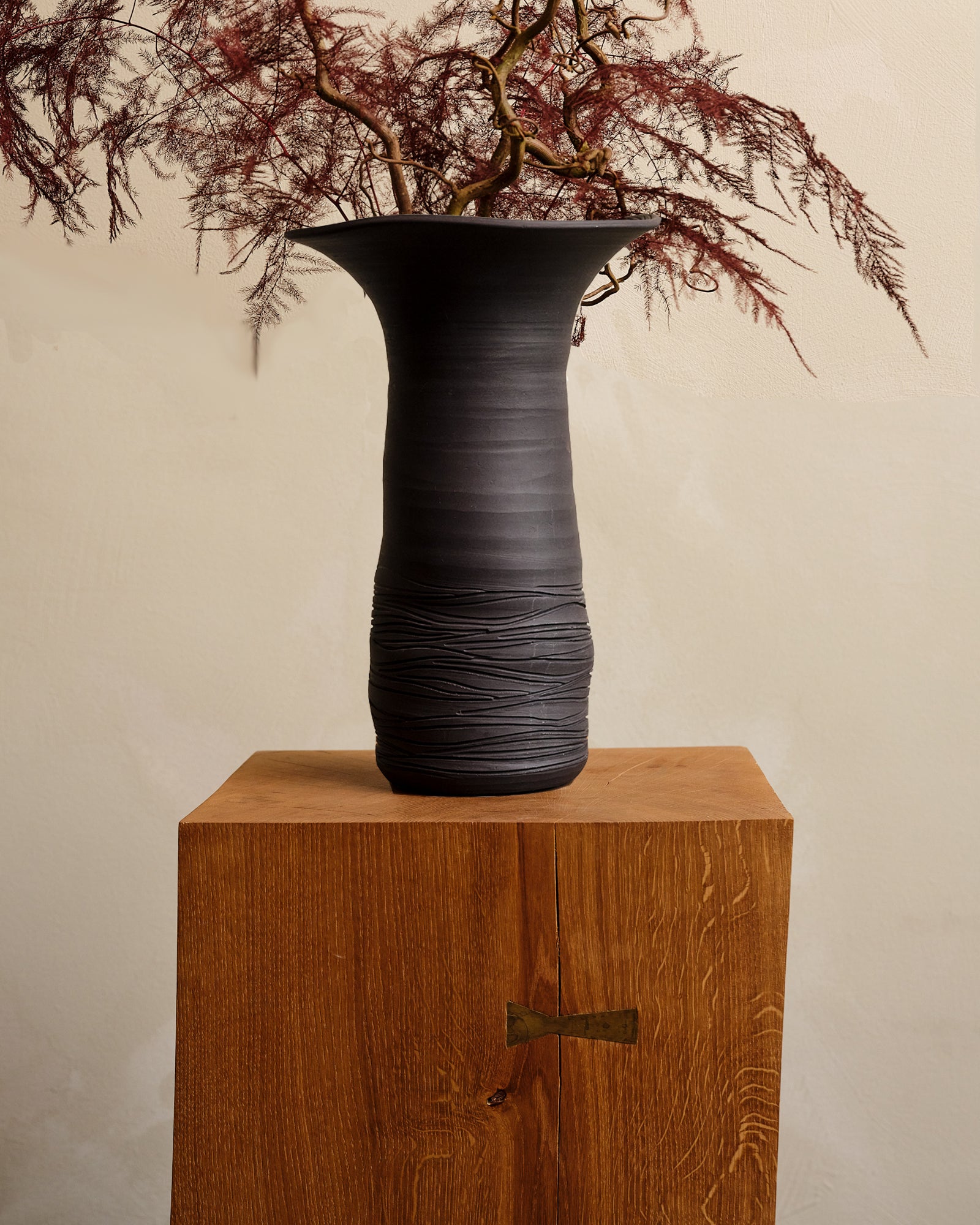 Black textured vase on a wooden pedestal with dried branches against a beige background