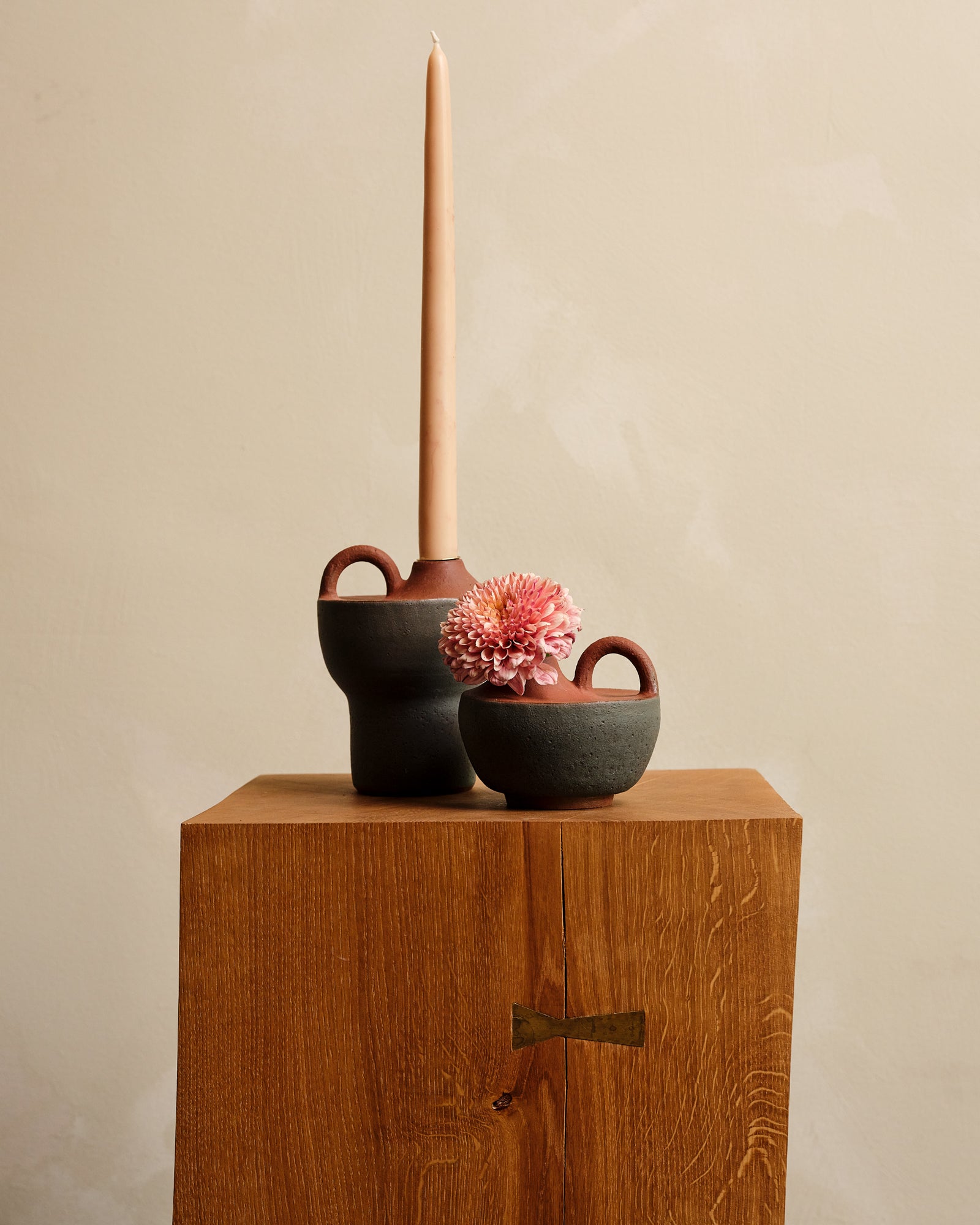 Two small ceramic vases with a pink flower and a tall candle on a wooden surface against a beige background