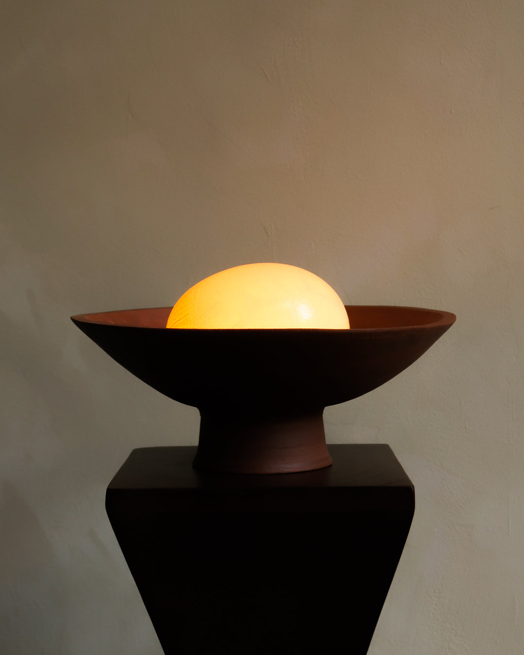 Decorative lamp with a glowing sphere inside a brown bowl on a dark pedestal against a plain background